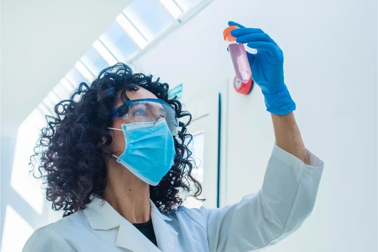 Woman in surgical mask and lab goggles holding up lab tube